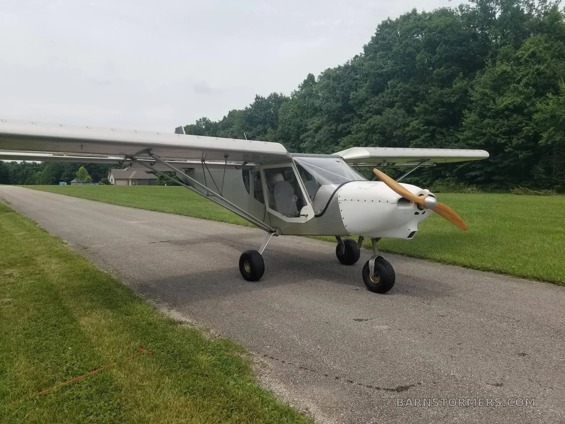 A light gray ultralight aircraft with a wooden propeller is parked on a paved path, with green grass and trees in the background.