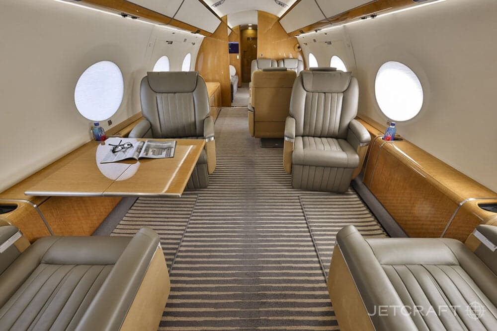 The luxurious cabin interior of a business jet featuring light brown leather seats, a wooden table, and striped carpet.
