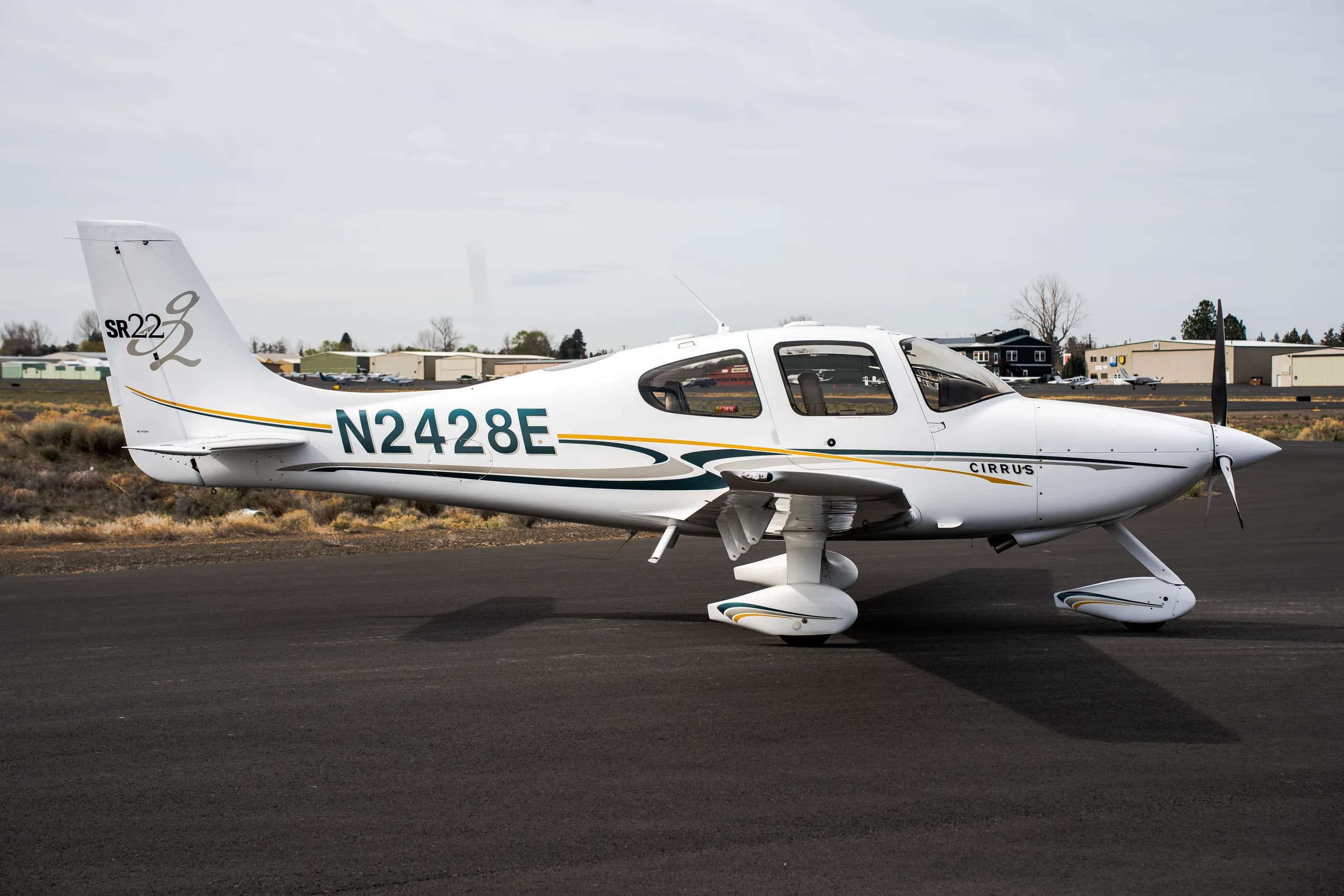 A side view of a white Cirrus SR22 aircraft with green and gold stripes, tail number N2428E, parked on an airfield.