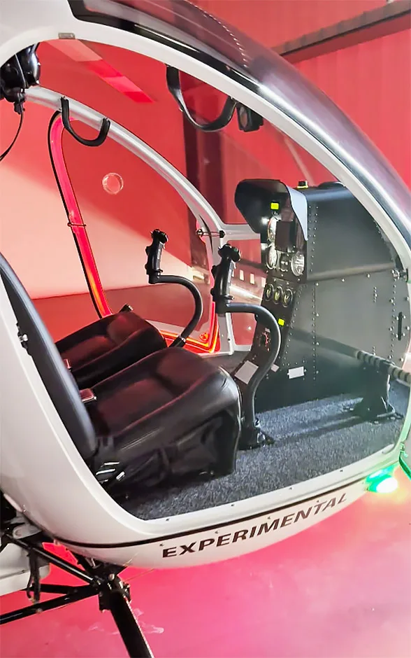 The cockpit of a white experimental helicopter with black seats and a minimalist instrument panel, illuminated by red light.