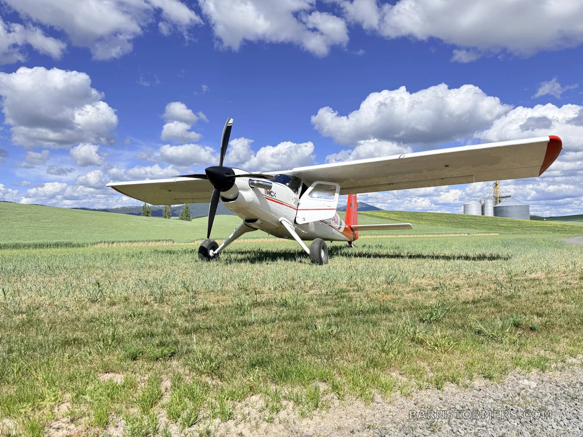 A white and red T-MCX aircraft (N634DV) is parked on a green field under a blue sky with white clouds, with silos in the distance.