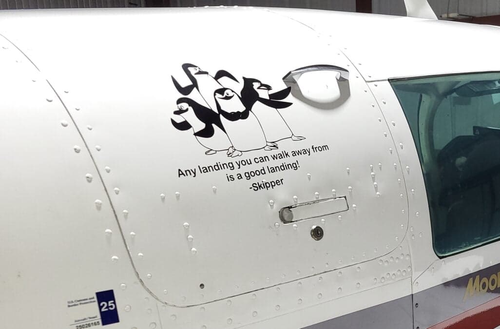 A close-up of the white fuselage of a Mooney M20J, showing a 'penguins' decal and the text 'Any landing you can walk away from is a good landing! -Skipper'.
