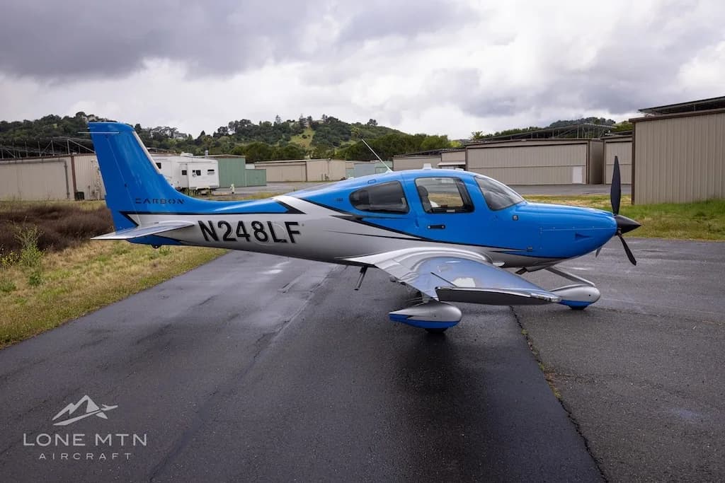 A blue and silver single-engine propeller aircraft (Cirrus SR22) with registration N248LF parked on a wet tarmac with hangars in the background.