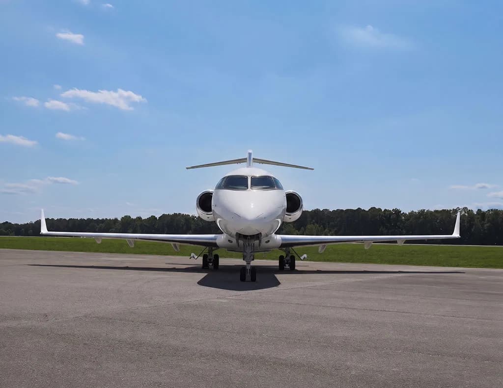 A white Bombardier Challenger 605 business jet seen from the front, parked on an airfield with trees in the background.
