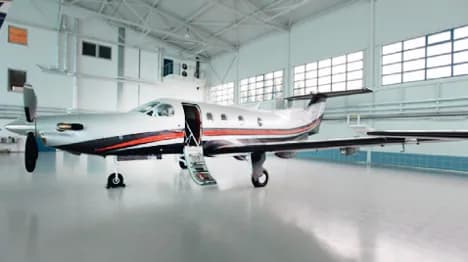 A silver Pilatus PC-12 turboprop aircraft with black and orange stripes is parked in a brightly lit hangar with its passenger door open.