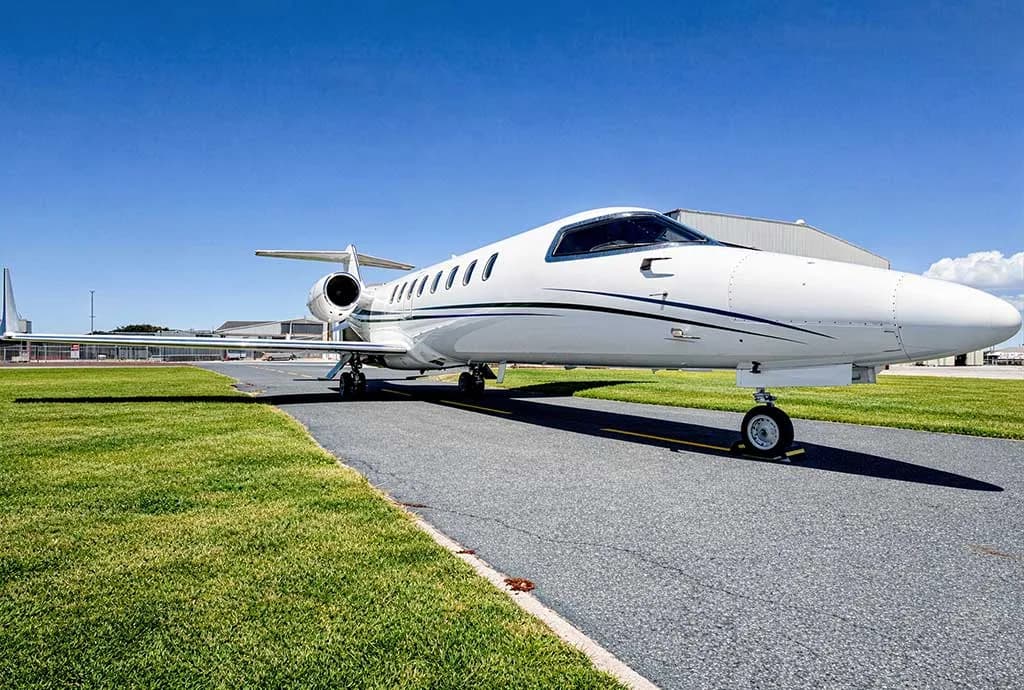 A white business jet with blue and black stripes is parked on an airport tarmac next to a grassy area, with hangars visible in the background.