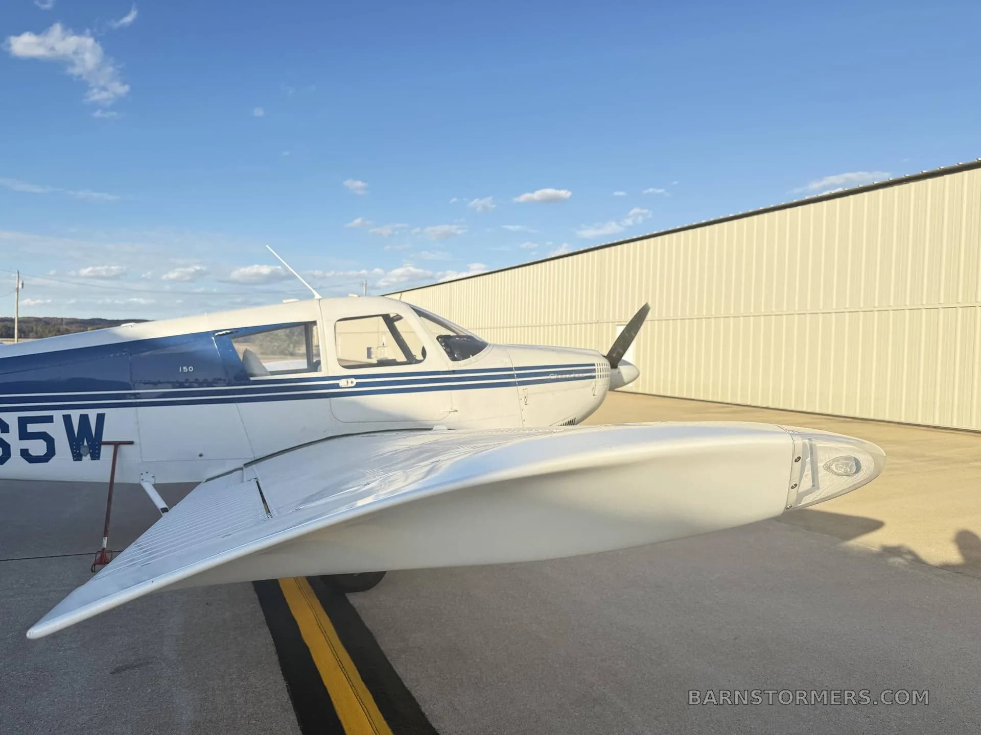 A white Piper Cherokee 150 aircraft with blue stripes parked on an tarmac next to a hangar under a blue sky.