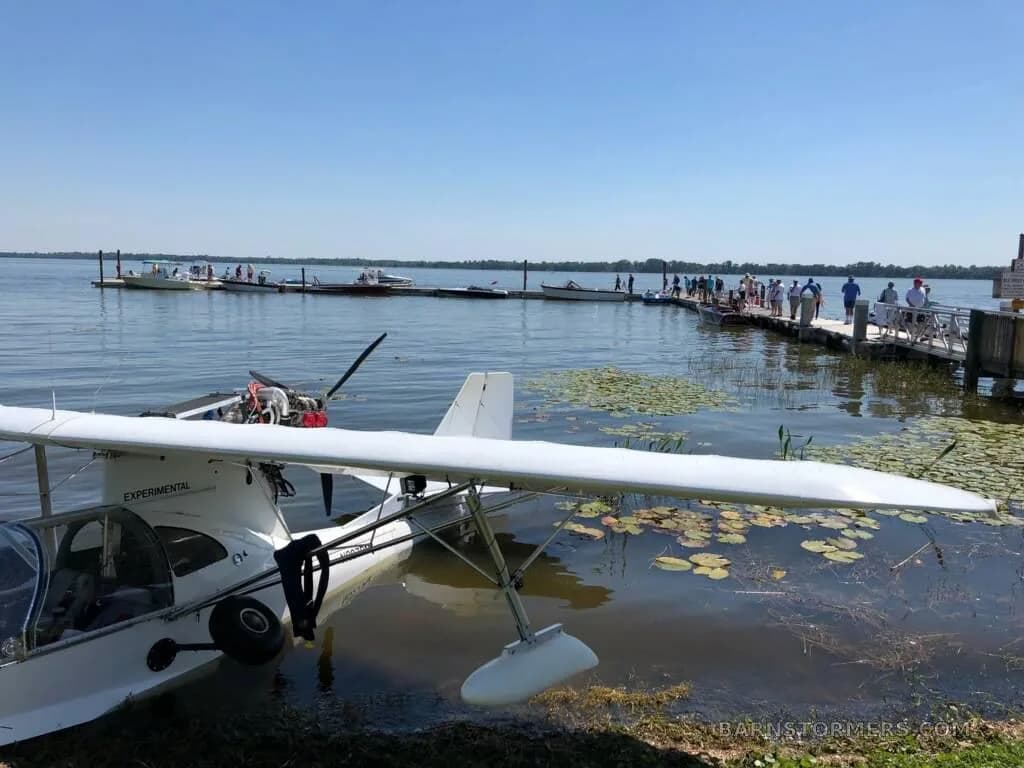 A white experimental seaplane is partially docked in water near a pier with people on a sunny day.