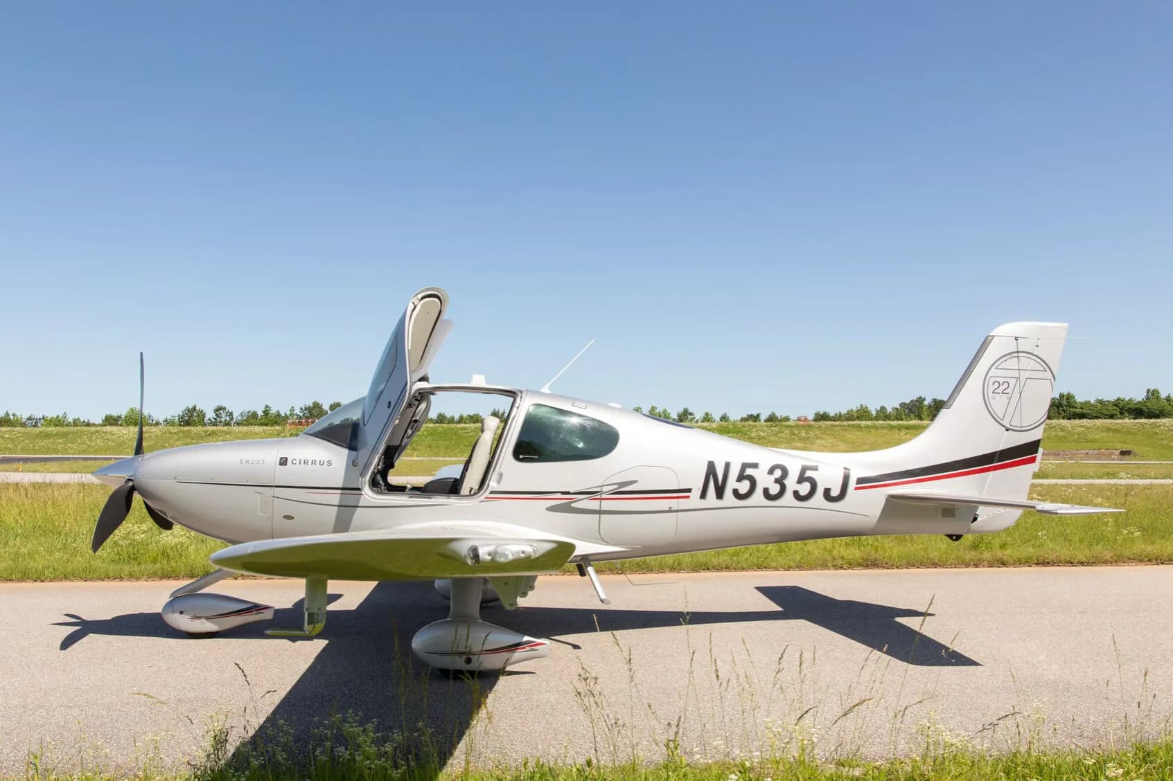 A silver Cirrus SR22T aircraft with registration N535J is parked with its doors open on an asphalt strip next to green grass under a clear blue sky.