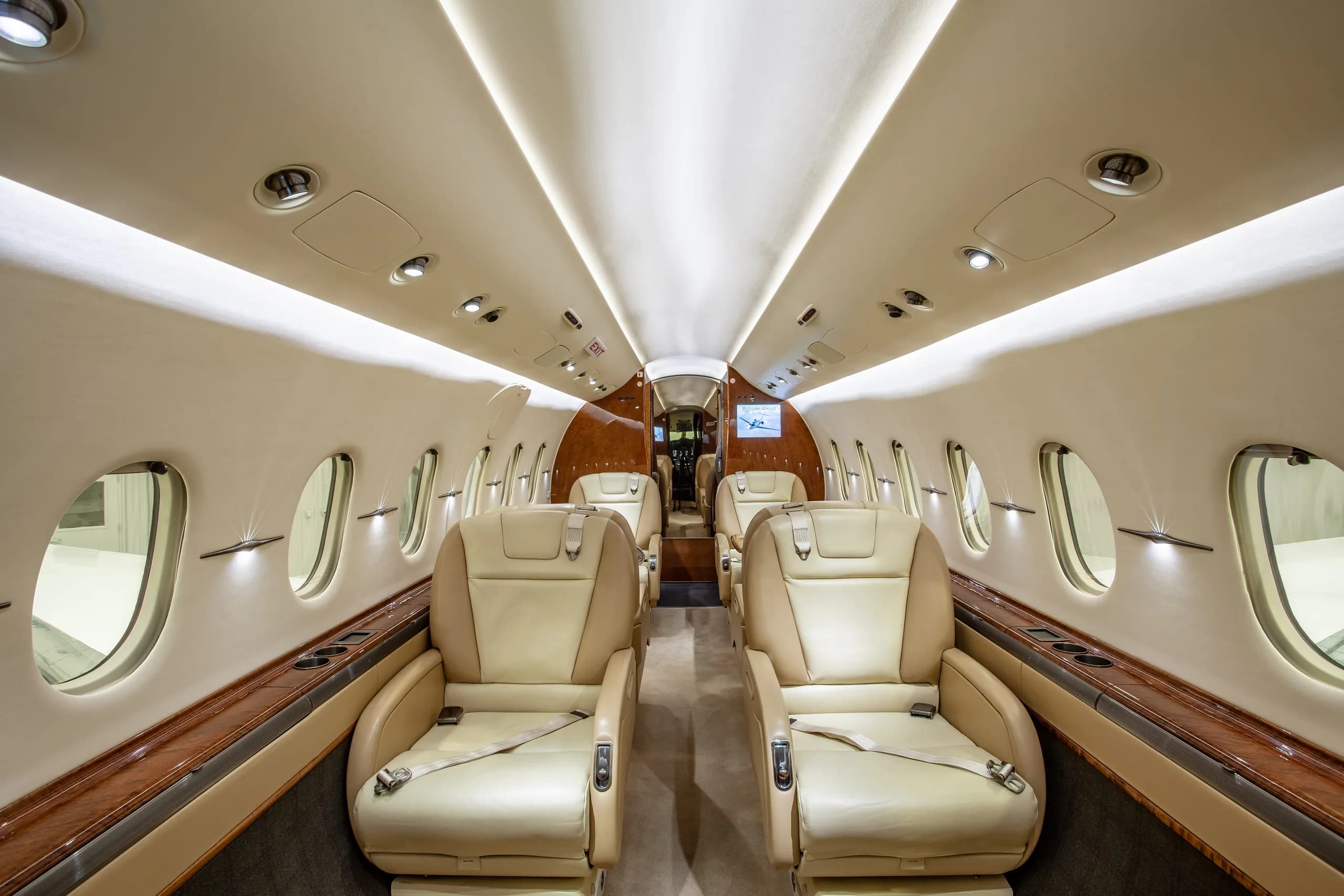A wide view of a private jet's interior cabin featuring beige leather seats, large windows, and wood trim.