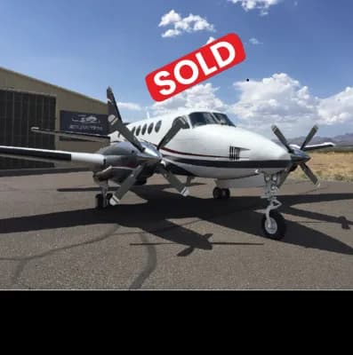 A white and dark-striped twin-engine turboprop aircraft, likely a King Air, parked on a tarmac next to a hangar.