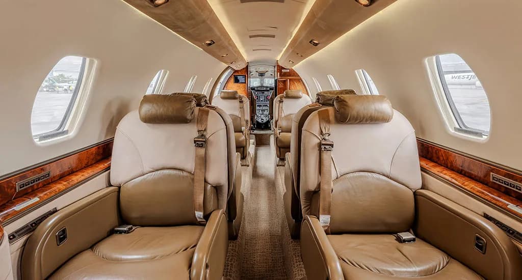A view down the aisle of a business jet's cabin, showing multiple tan and brown leather seats and overhead lighting.