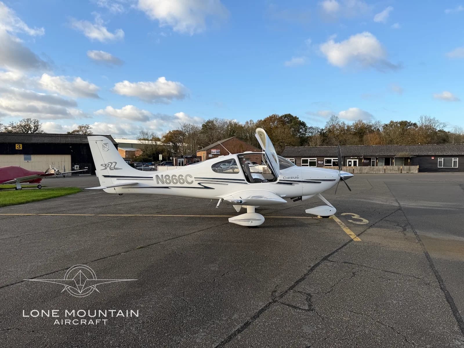 A side view of a white Cirrus SR22 aircraft with black and grey stripes, registration N866C, parked on an asphalt apron with its doors open.