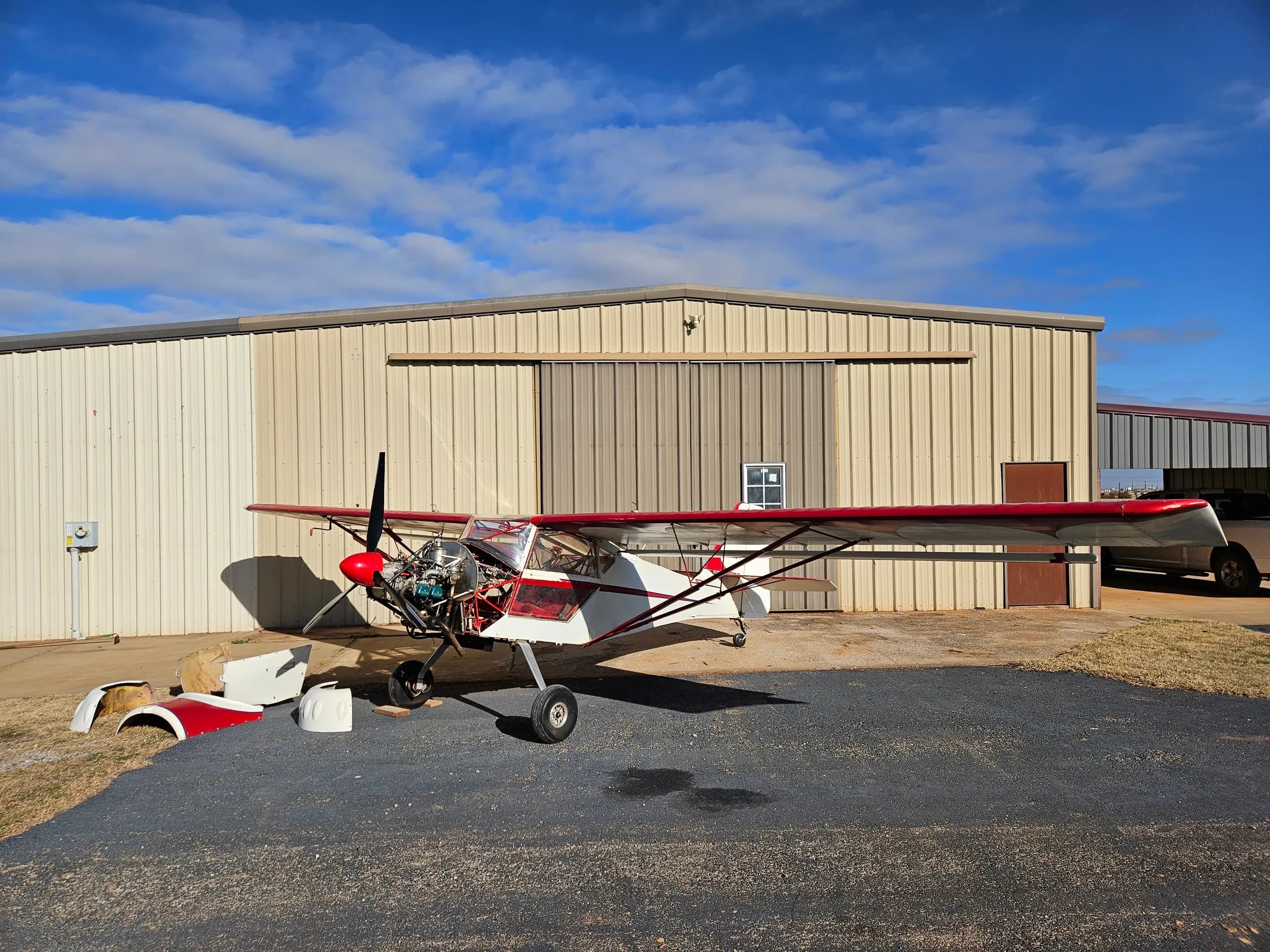 A white and red experimental aircraft with its engine exposed, parked on an asphalt apron in front of a large hangar.