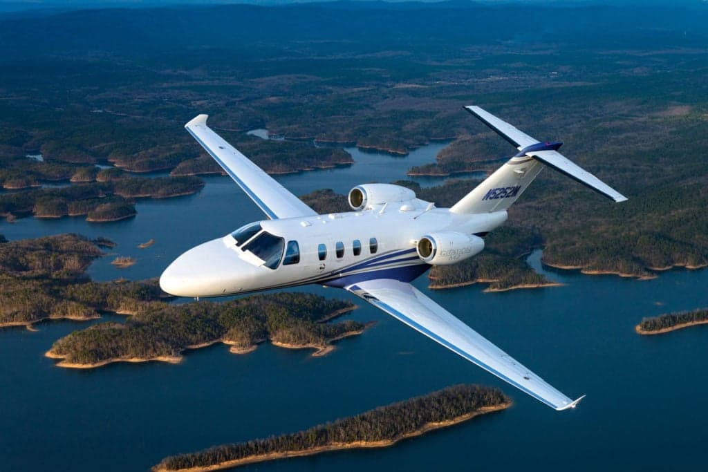 A Citation M2 aircraft in flight over a lake landscape.