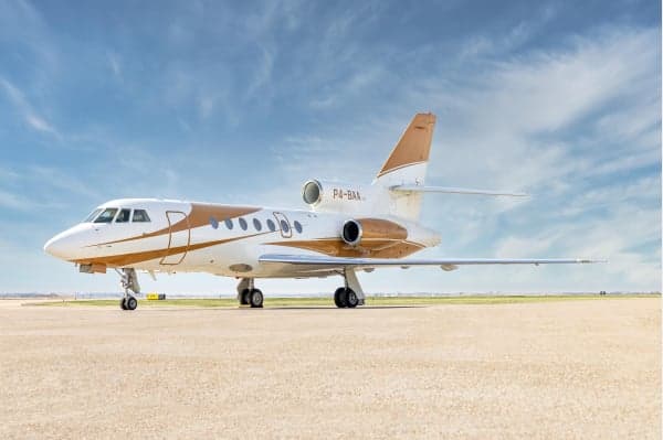 A Dassault Falcon 900 business jet with a white and gold paint scheme is parked on an airfield under a blue sky.