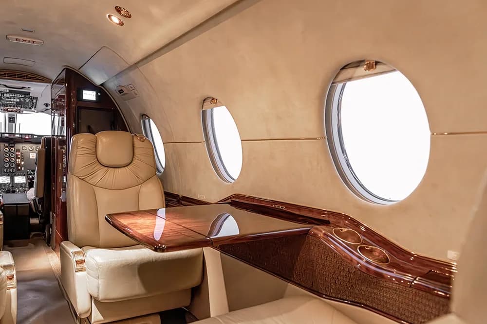View inside a private jet cabin showing a light-colored leather seat, a polished wood table, and multiple windows.