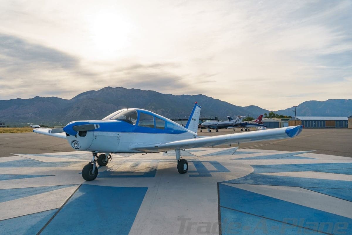 A front three-quarter view of the blue and white single-engine aircraft N8718N on a patterned tarmac with other planes and mountains in the background.
