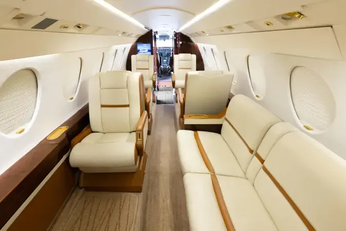 A view down the private jet cabin aisle, showing cream leather seating, a sofa, and the cockpit entrance in the distance.