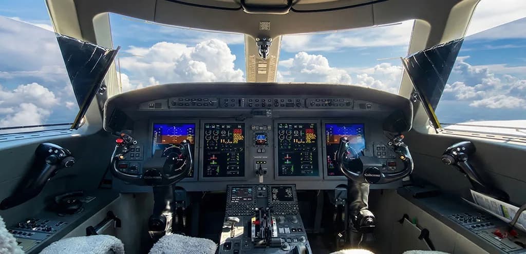The cockpit of the private jet features multiple display screens and yokes, with clouds visible through the windshield.