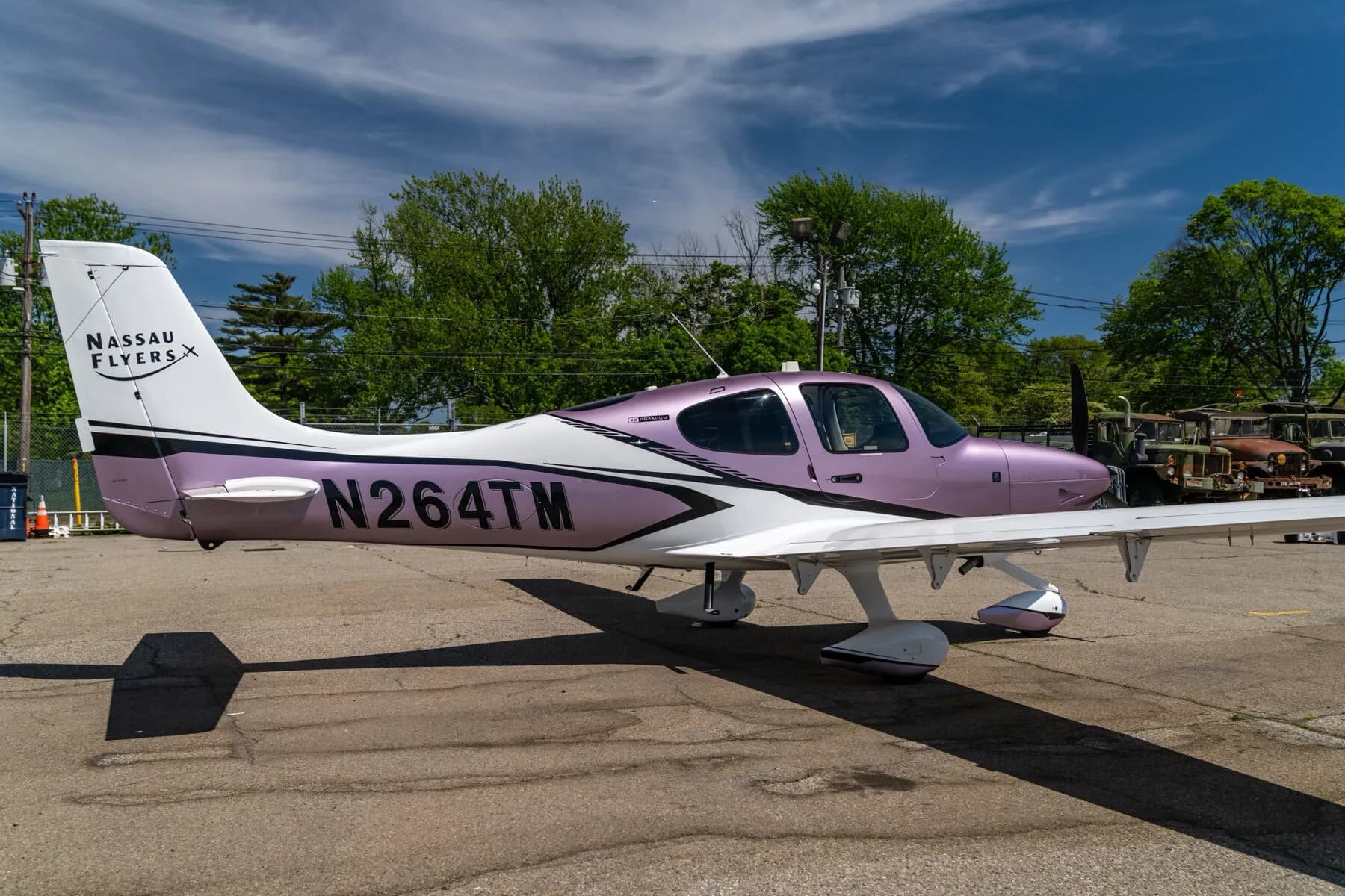 A Cirrus SR22 aircraft with a metallic lavender and white paint scheme, registration N264TM, parked on an airfield with trees and old vehicles in the background.