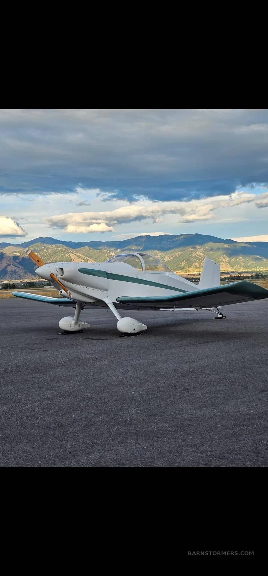 A white single-engine aircraft with teal stripes is parked on an airfield with mountains and a cloudy sky in the background.