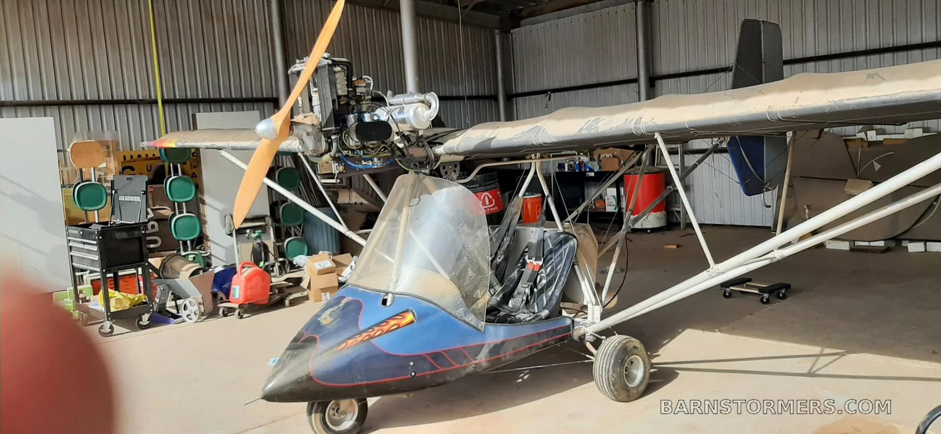 A blue and black ultralight aircraft with an exposed engine and propeller, parked inside a dusty hangar.