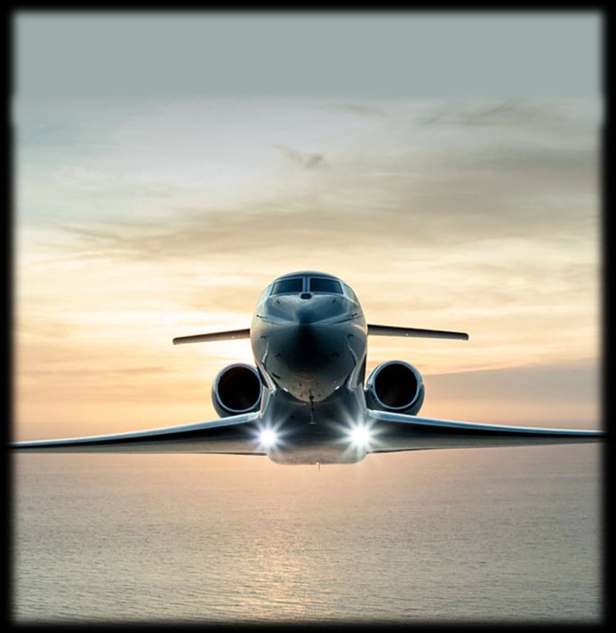 Front view of a private jet flying over the ocean at sunset.