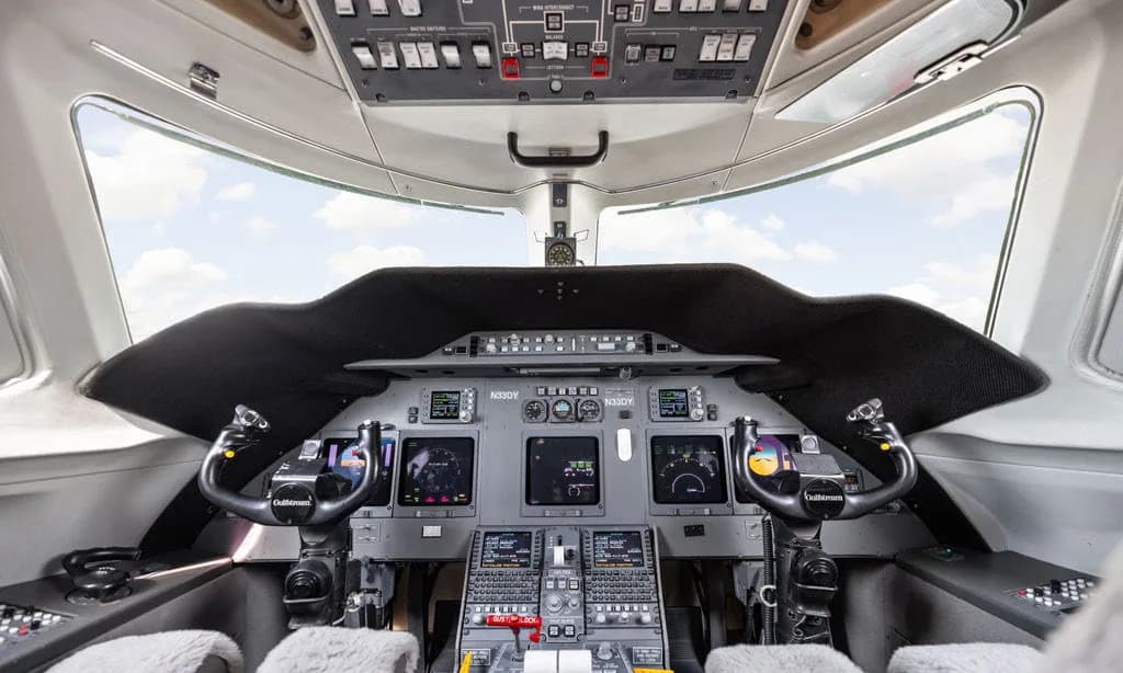 The cockpit of a private jet features multiple digital displays, control yokes, and an overhead panel.