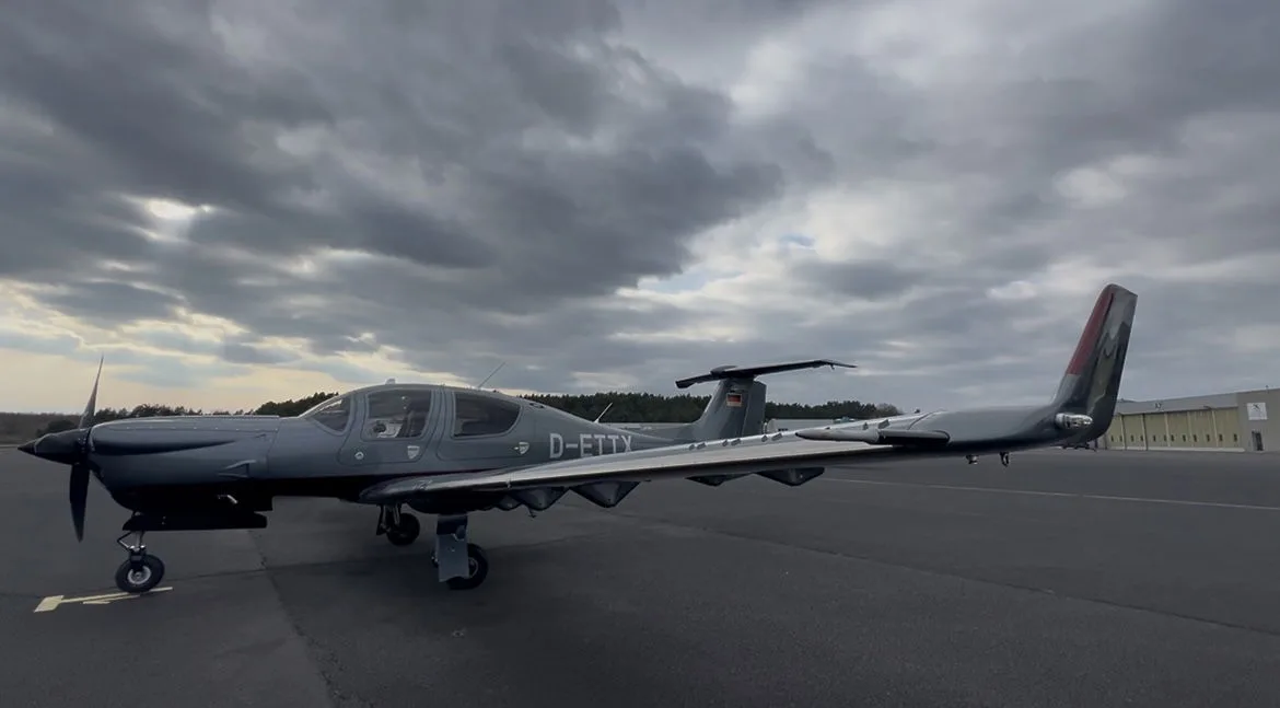 A gray Diamond DA50 RG aircraft is parked on the tarmac under a cloudy sky.