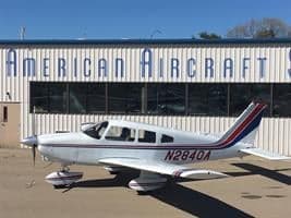A white single-engine Piper aircraft with red and blue stripes, tail number N2840A, parked on an airfield in front of a building labeled 'AMERICAN AIRCRAFT'.