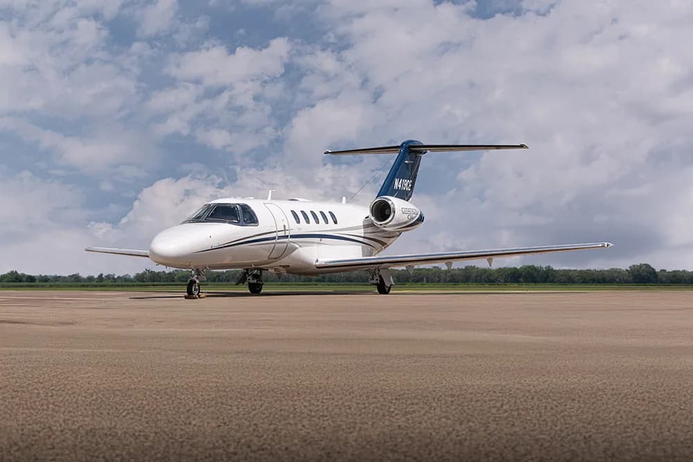 A white private jet with blue and grey stripes on the tail and fuselage, parked on an airfield under a cloudy sky.