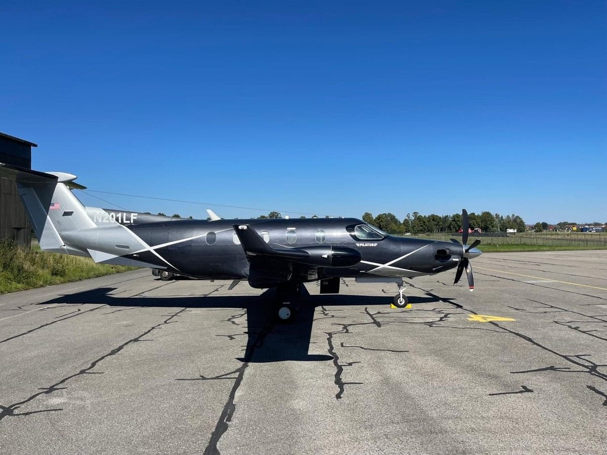 A dark grey and silver Pilatus PC-12 turboprop aircraft with white accents is parked on an asphalt tarmac, viewed from the front-left.