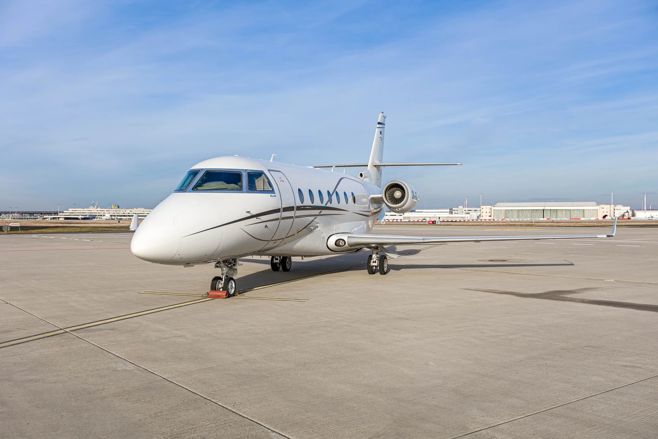 A white Gulfstream G200 business jet with brown accent stripes parked on an airport tarmac, viewed from the front-right.