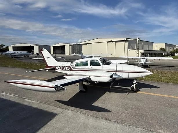 A white twin-engine aircraft with N87393 on the tail, parked on an airfield with hangars.