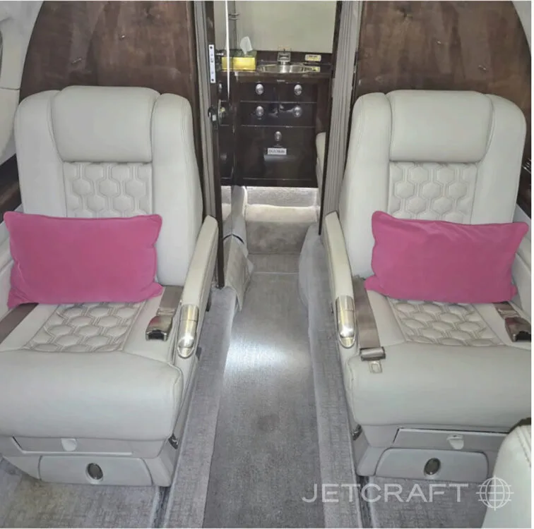The interior cabin of a business jet, showing two cream-colored seats with pink pillows and a galley area in the background.