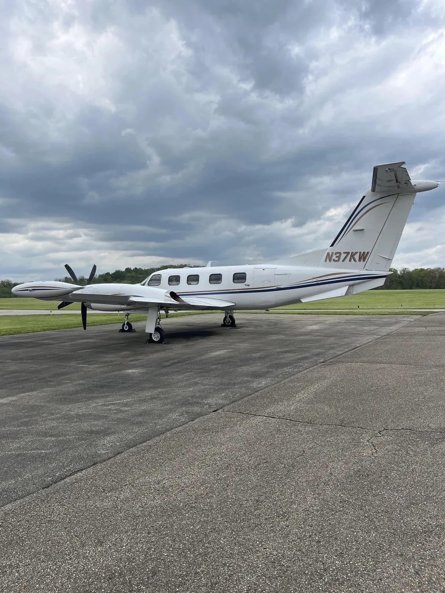 Side view of a white twin-engine turboprop aircraft with blue and gold stripes on the tail (N37KW), parked on an airfield.