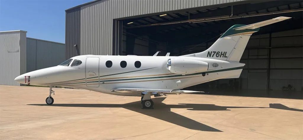 A white Beechcraft Premier 1 private jet, registration N76HL, with green and gold stripes, parked on a concrete tarmac next to an open hangar.