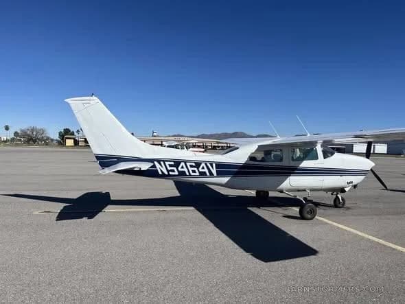 A white Cessna 182 with blue stripes, registration N5464V, parked on an asphalt ramp under a clear sky.