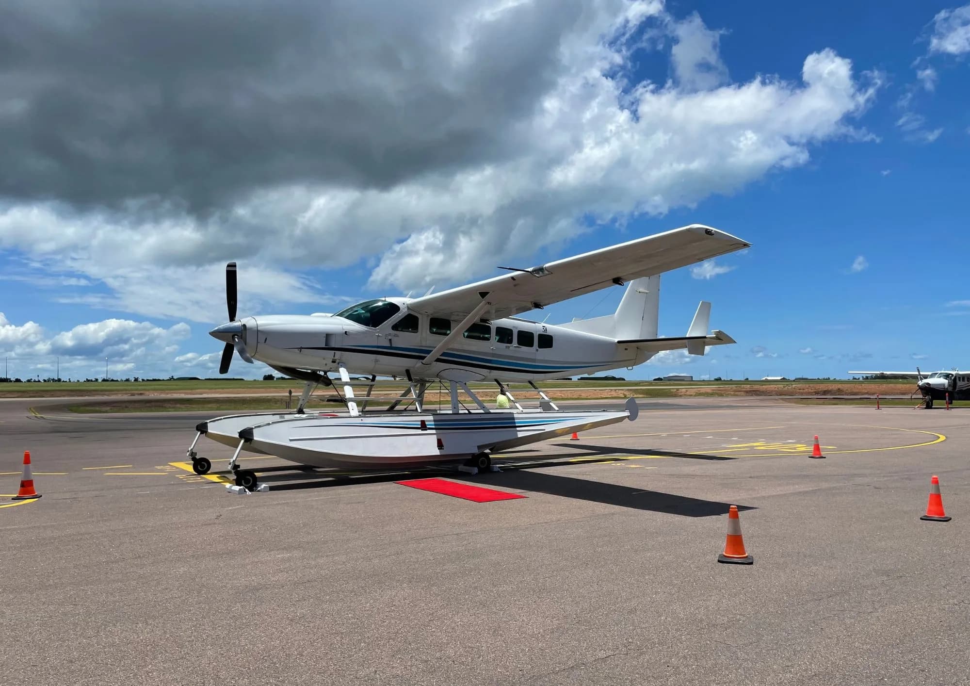 A white single-engine turboprop seaplane with blue stripes is parked on an asphalt tarmac under a partly cloudy sky.