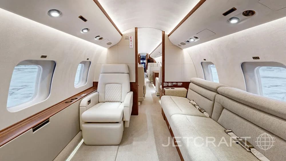 A view down the aisle of a business jet cabin, showing cream-colored seats and a sofa with wood accents.