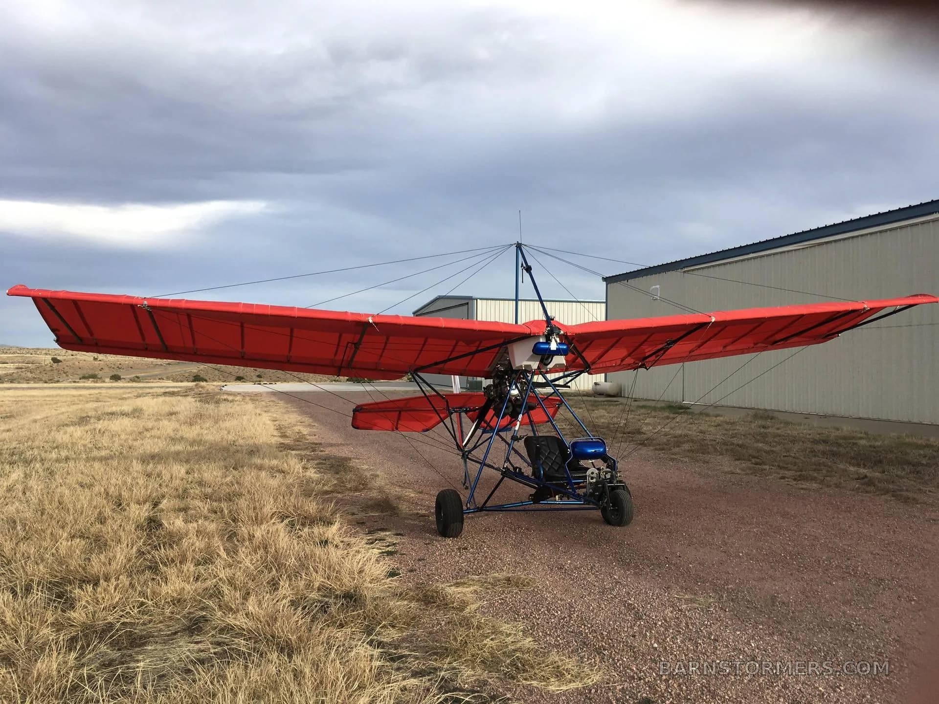 A red ultralight aircraft with a blue frame, parked on a dirt path in front of hangars.