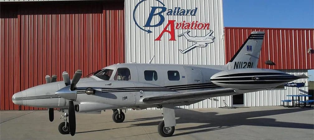Twin-engine aircraft parked in front of a hangar with the 'Ballard Aviation' logo.