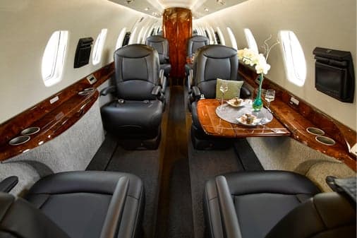The luxurious interior of a private jet cabin featuring black leather seats, polished wood accents, and a set table.