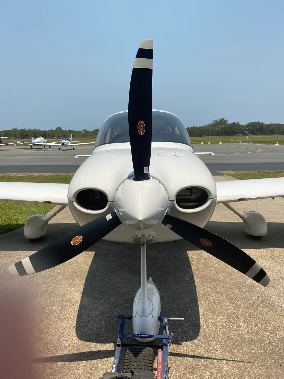 A white single-engine propeller aircraft is parked on an airfield, viewed from the front with other planes in the background.