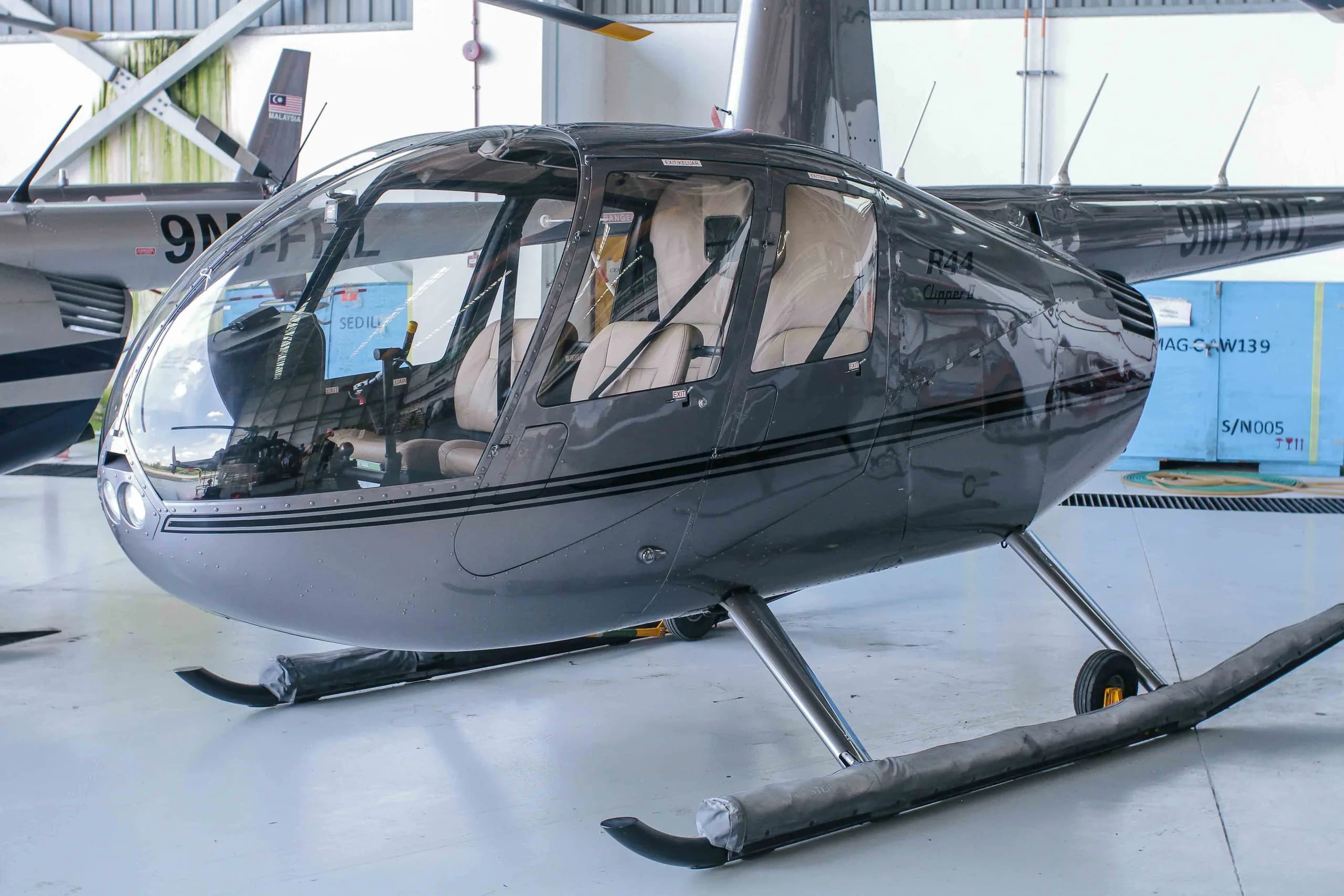 A dark grey Robinson R44 Clipper II helicopter with black stripes is parked inside a hangar.