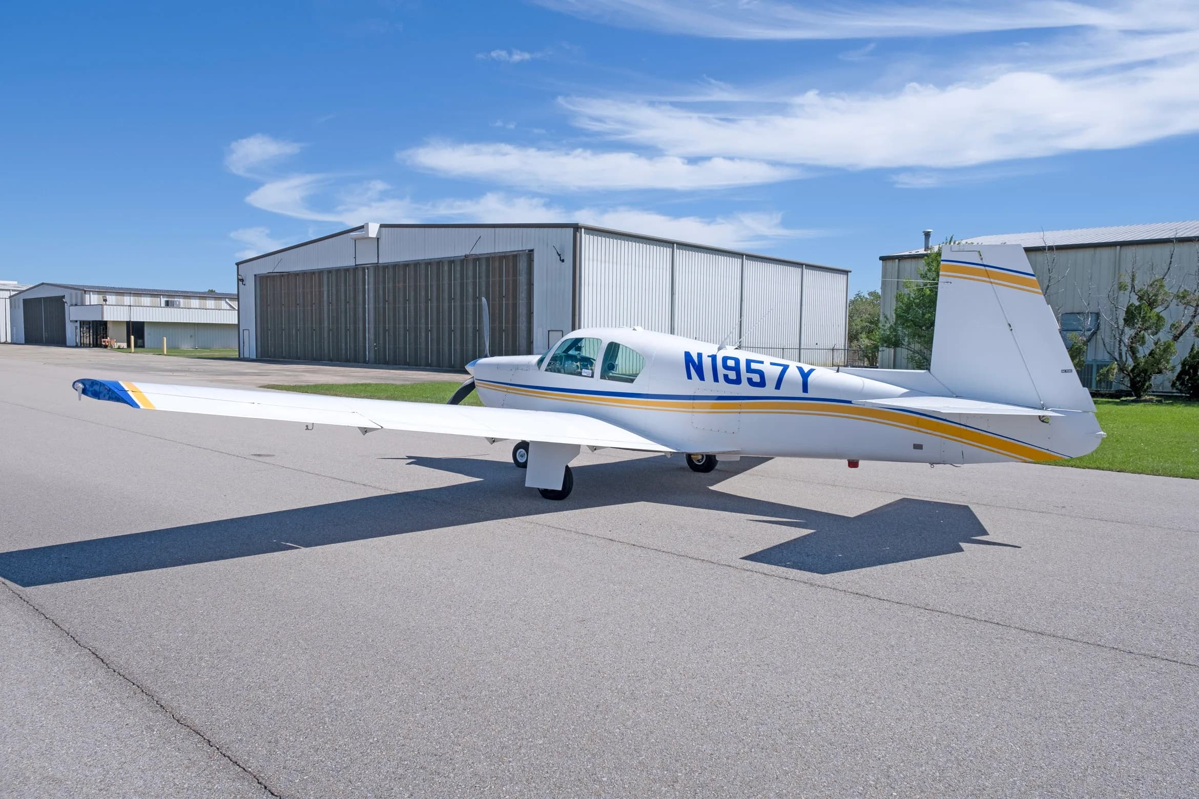 A rear-side view of a white single-engine propeller aircraft with blue and yellow stripes, registration N1957Y, parked on an asphalt apron.