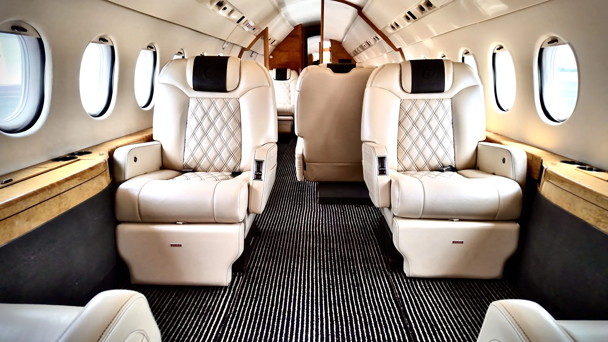 The luxurious interior cabin of a private jet, featuring cream-colored quilted leather seats and wood trim.
