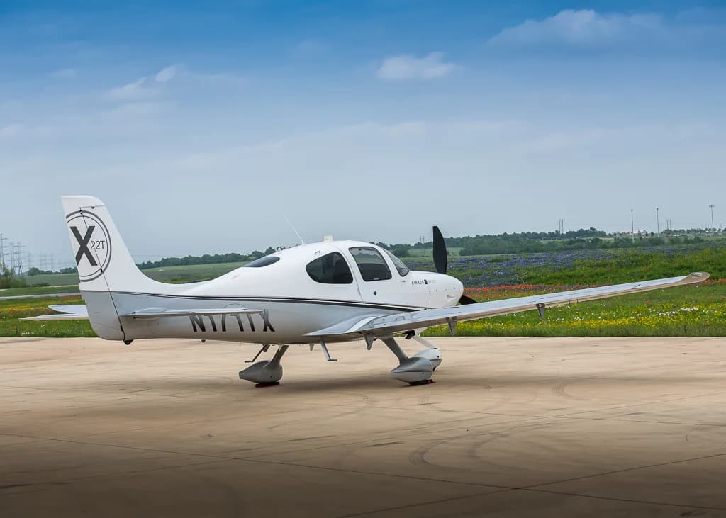 A side view of the white and silver Cirrus SR22T aircraft, N171TX, parked on a tarmac next to a field of wildflowers.