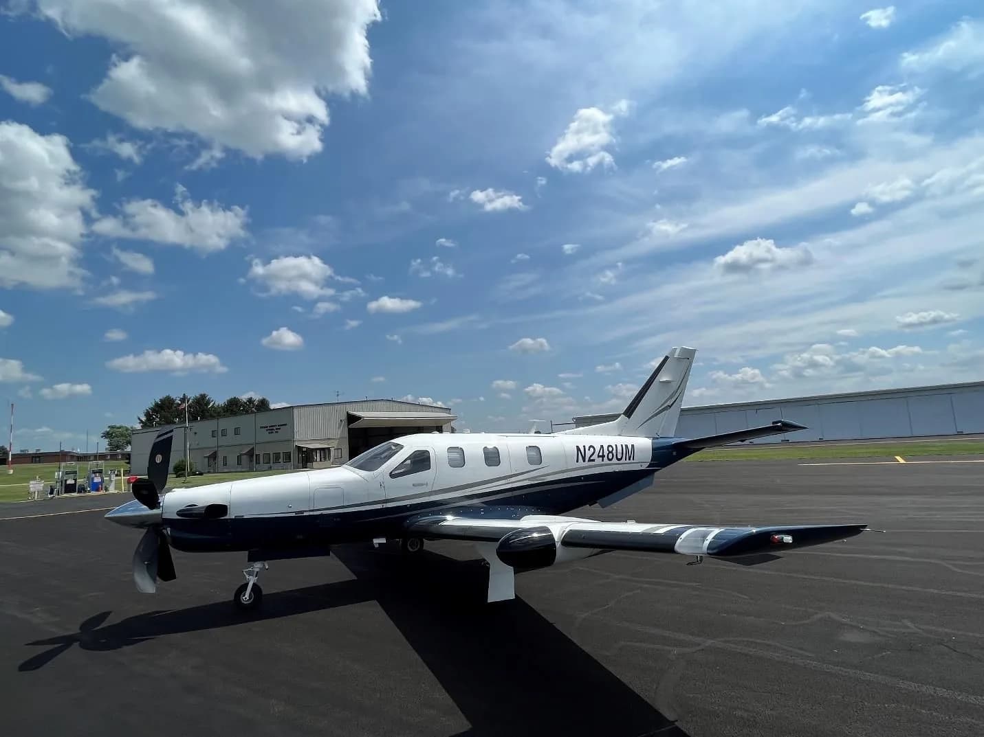 A three-quarter front view of the N248UM aircraft, a white and blue turboprop, parked on an airfield with buildings in the background.